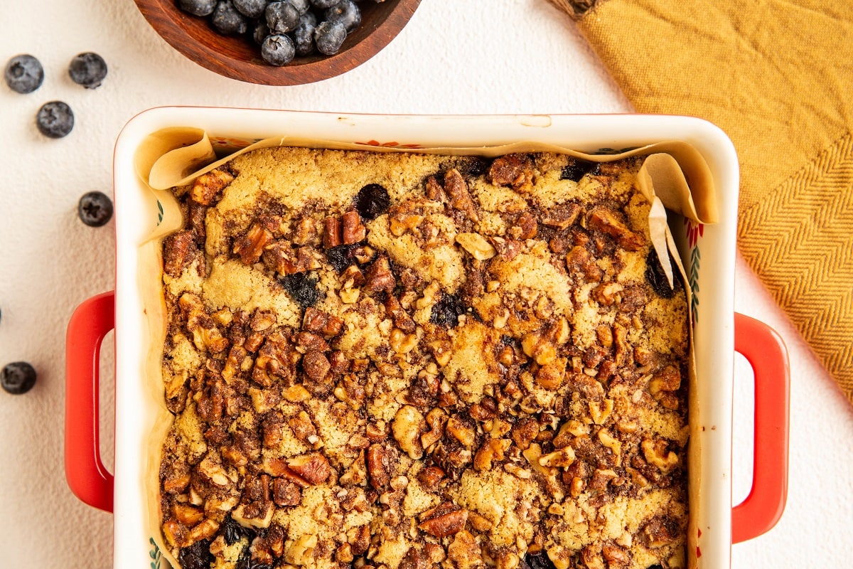 finished blueberry coffee cake in a cake pan with a golden napkin to the side and blueberries all around.