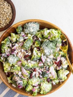 Wooden bowl full of broccoli salad with a striped napkin to the side and a bowl of sunflower seeds