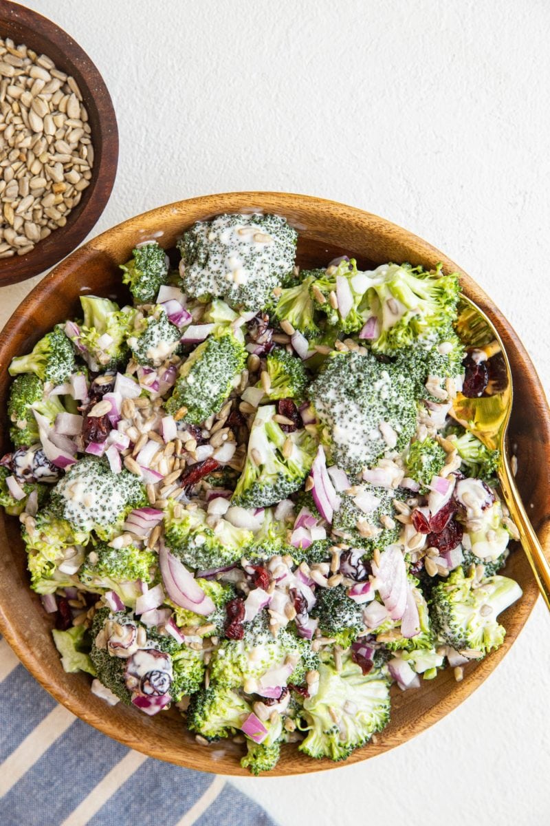 Wooden bowl full of broccoli salad with a striped napkin to the side and a bowl of sunflower seeds