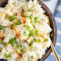 wooden bowl of potato salad with green onions on top and a spoon. Blue napkin off the the side and pepper grinder.