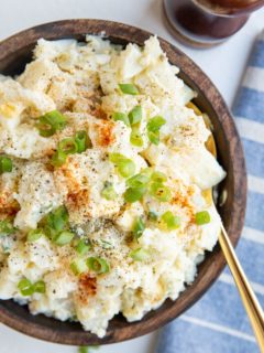 wooden bowl of potato salad with green onions on top and a spoon. Blue napkin off the the side and pepper grinder.