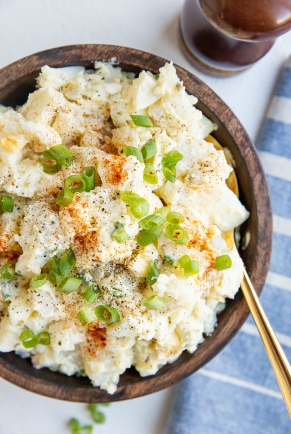 wooden bowl of potato salad with green onions on top and a spoon. Blue napkin off the the side and pepper grinder.