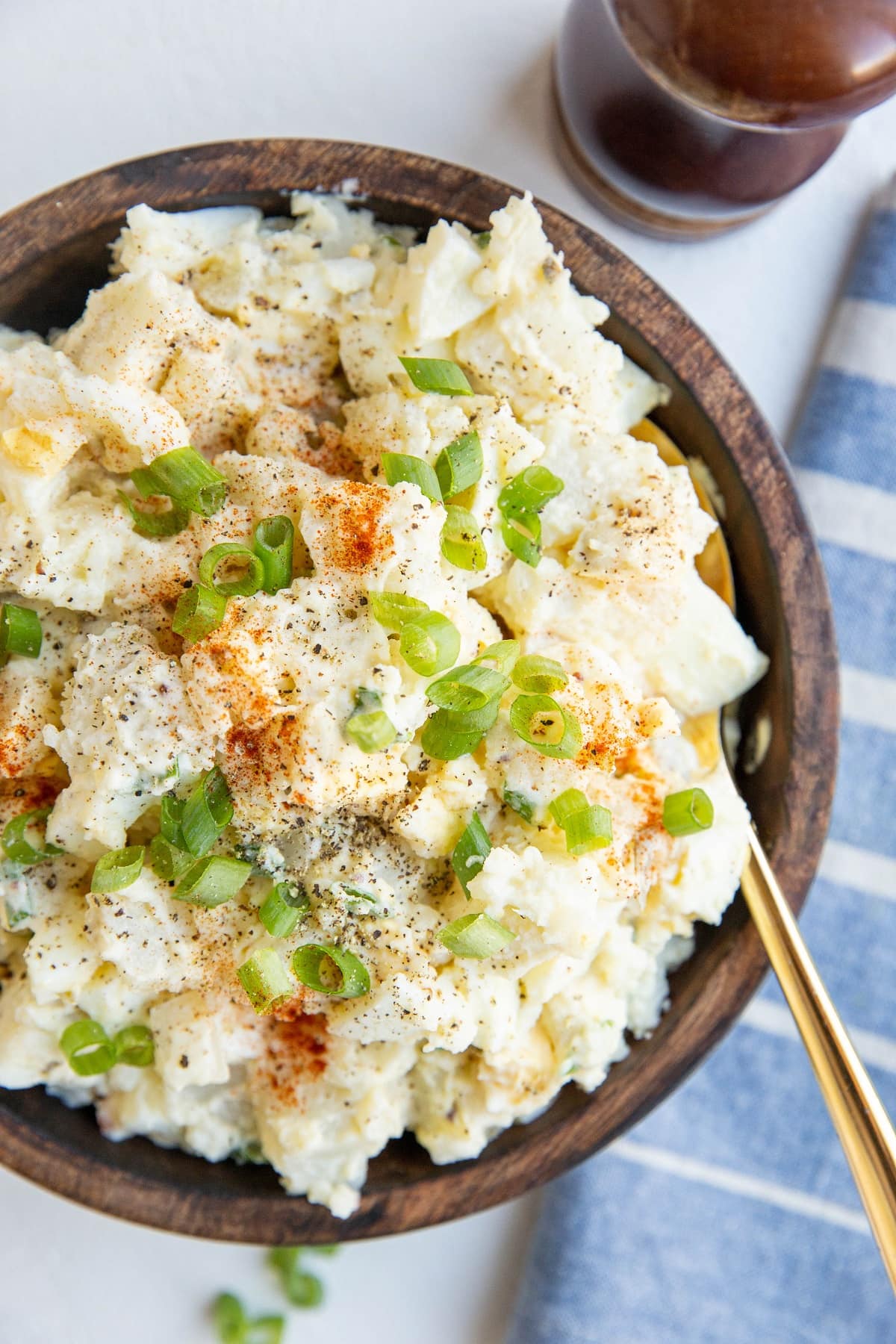 wooden bowl of potato salad with green onions on top and a spoon. Blue napkin off the the side and pepper grinder.