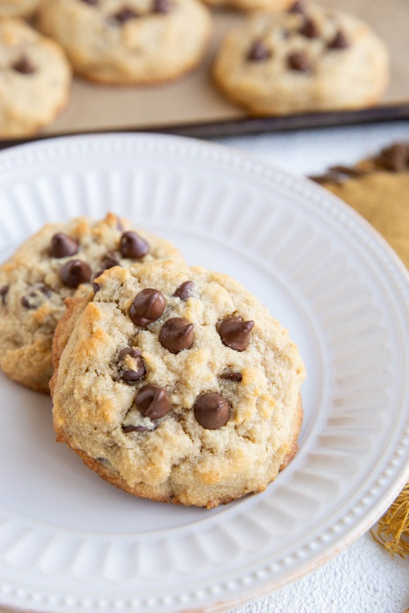 White plate with two coconut flour cookies on top with tray of cookies in the background.