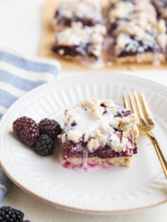 White plate with a blackberry crumb bar and fresh blackberries to the side and a gold fork.