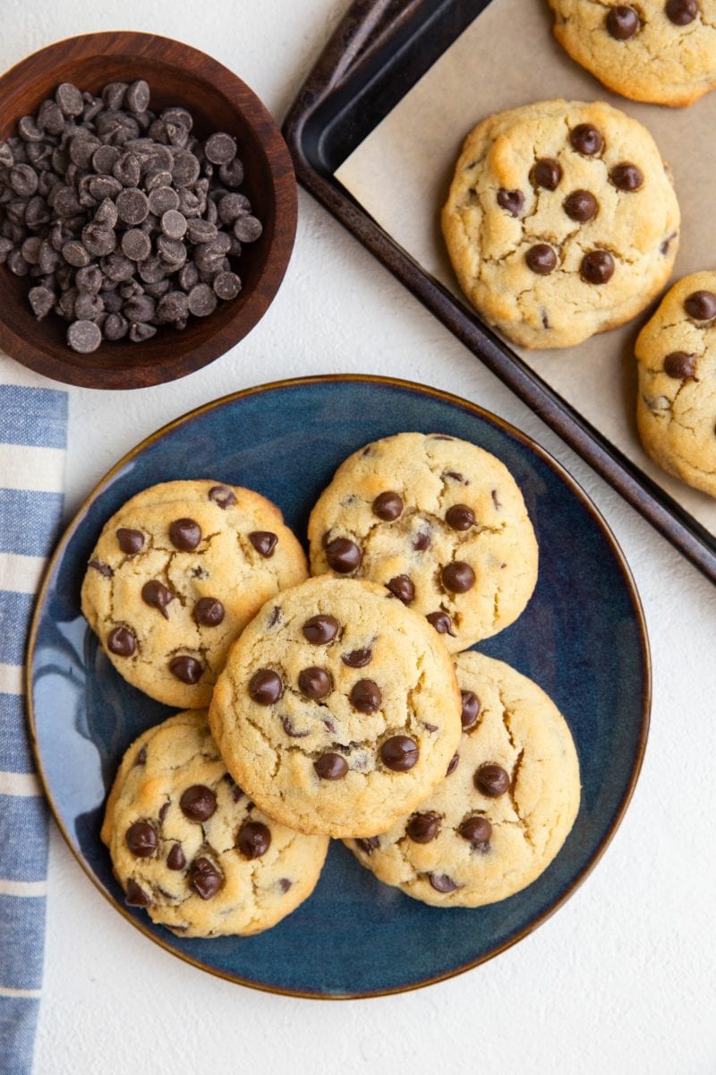 blue plate of chocolate chip cookies with a cookie sheet of cookies and a bowl of chocolate chips