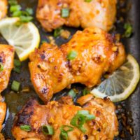 Casserole dish with baked boneless lemon garlic chicken thighs with sliced of lemon and green onion.