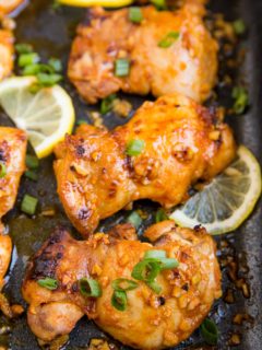 Casserole dish with baked boneless lemon garlic chicken thighs with sliced of lemon and green onion.