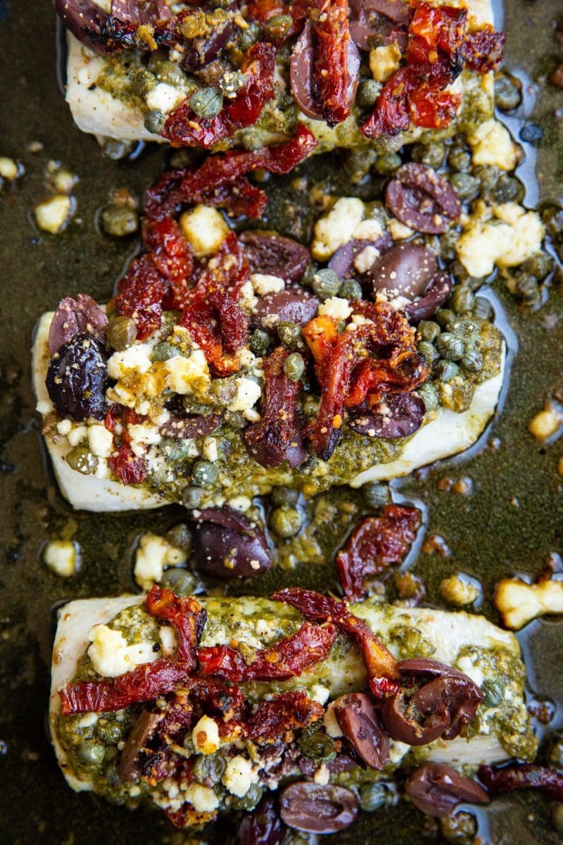 Baked cod recipe with pesto sauce on top and sun-dried tomatoes in a casserole dish.