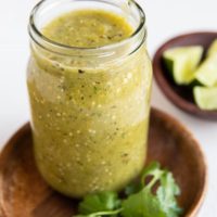 Jar of green tomatillo salsa on a wooden plate with bowl of limes in the background