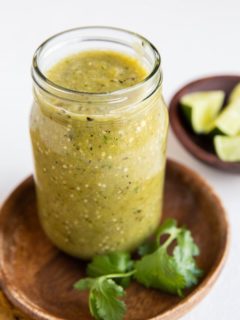 Jar of green tomatillo salsa on a wooden plate with bowl of limes in the background