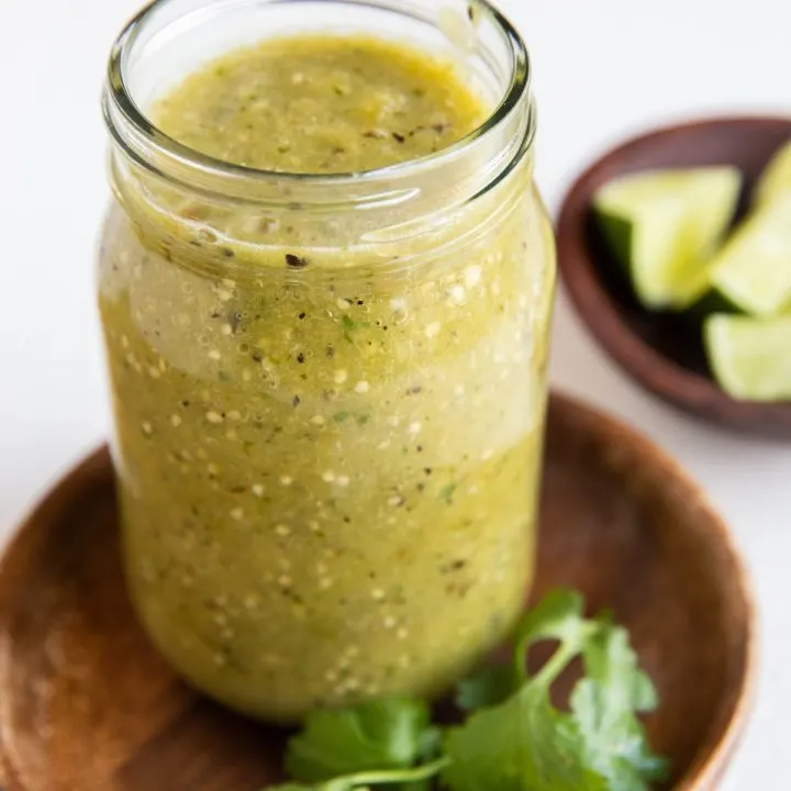 Jar of green tomatillo salsa on a wooden plate with bowl of limes in the background