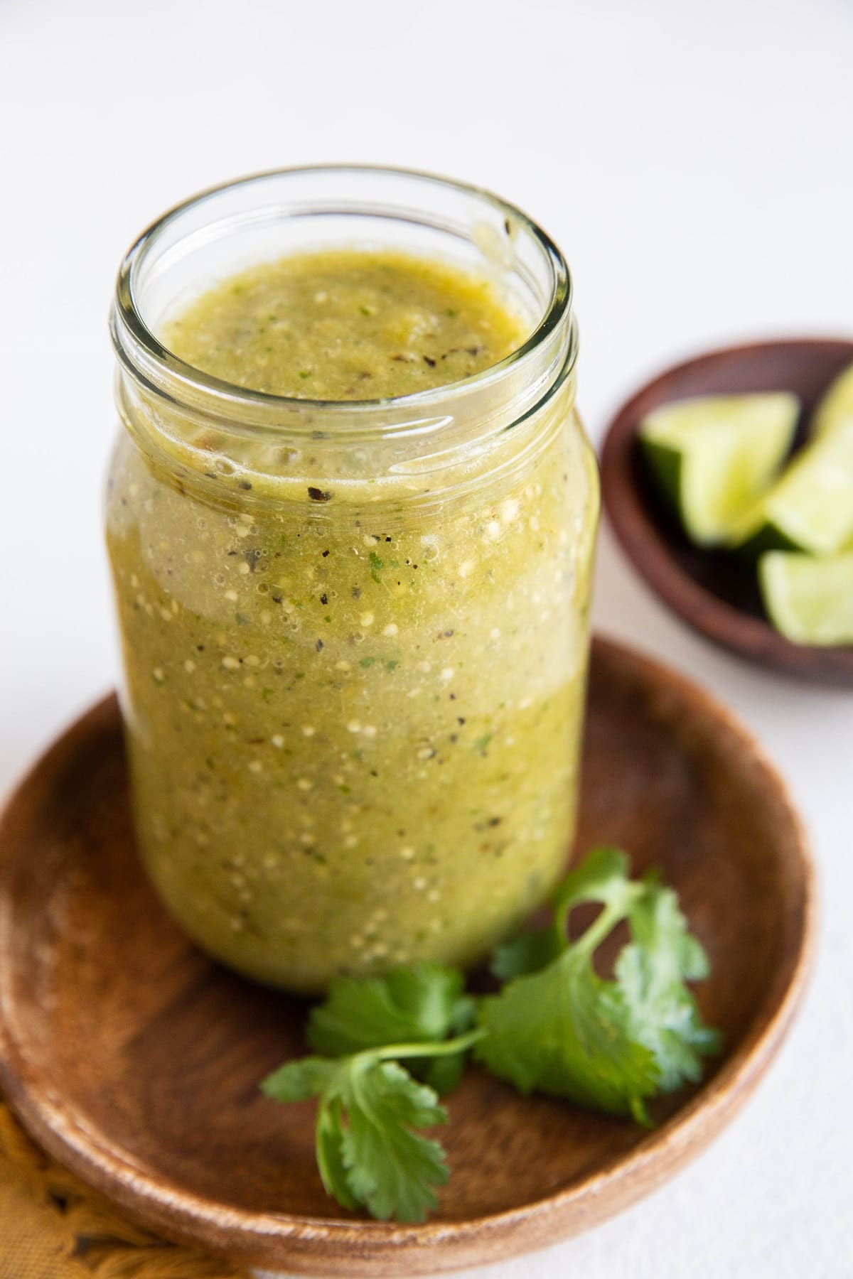 Jar of green tomatillo salsa on a wooden plate with bowl of limes in the background