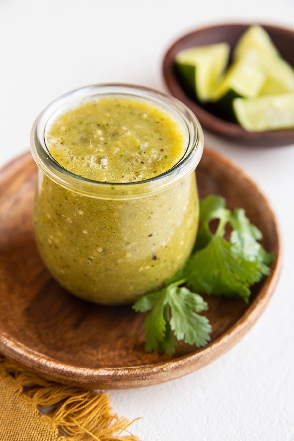 small jar of salsa verde sitting on a wooden plate with cilantro to the side and limes in a bowl