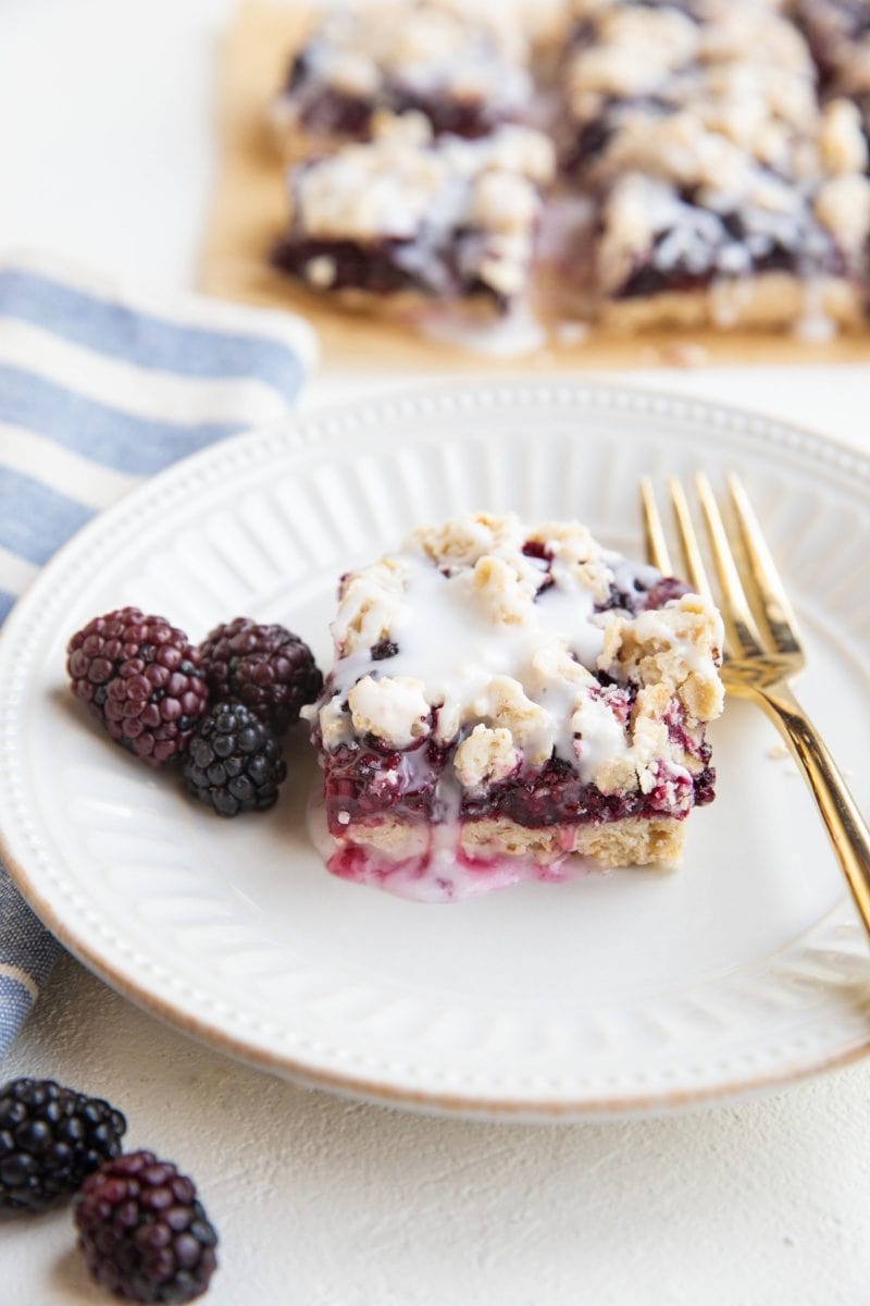 White plate with a blackberry crumb bar and fresh blackberries to the side and a gold fork.