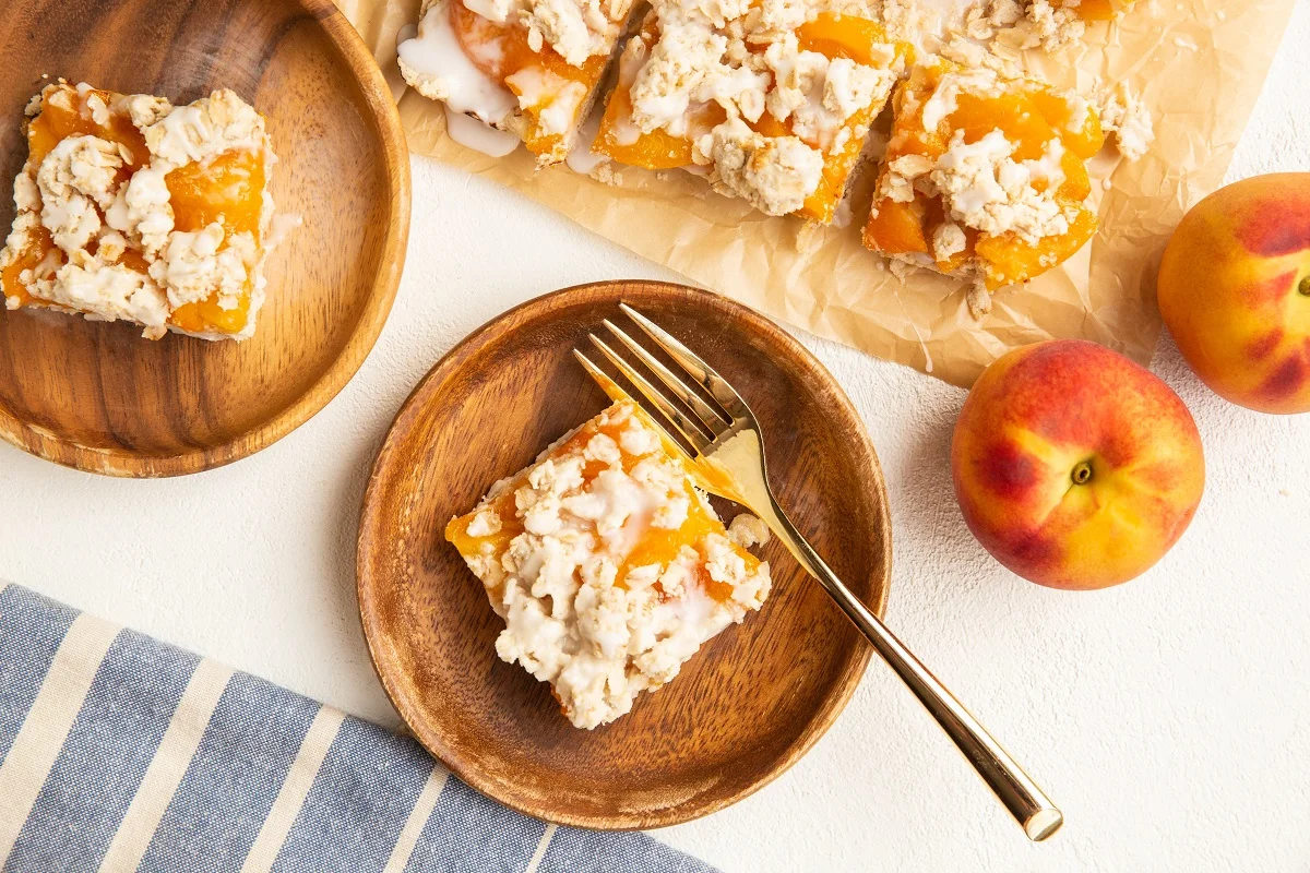 top down image of two wooden plates with slices of peach pie bars. Blue striped napkin and fresh peaches to the side.