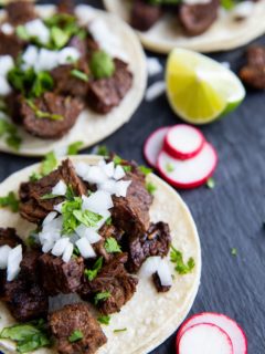 three carne asada tacos on a black slate with slices of turnips and limes to the sides