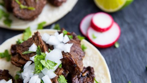 three carne asada tacos on a black slate with slices of turnips and limes to the sides