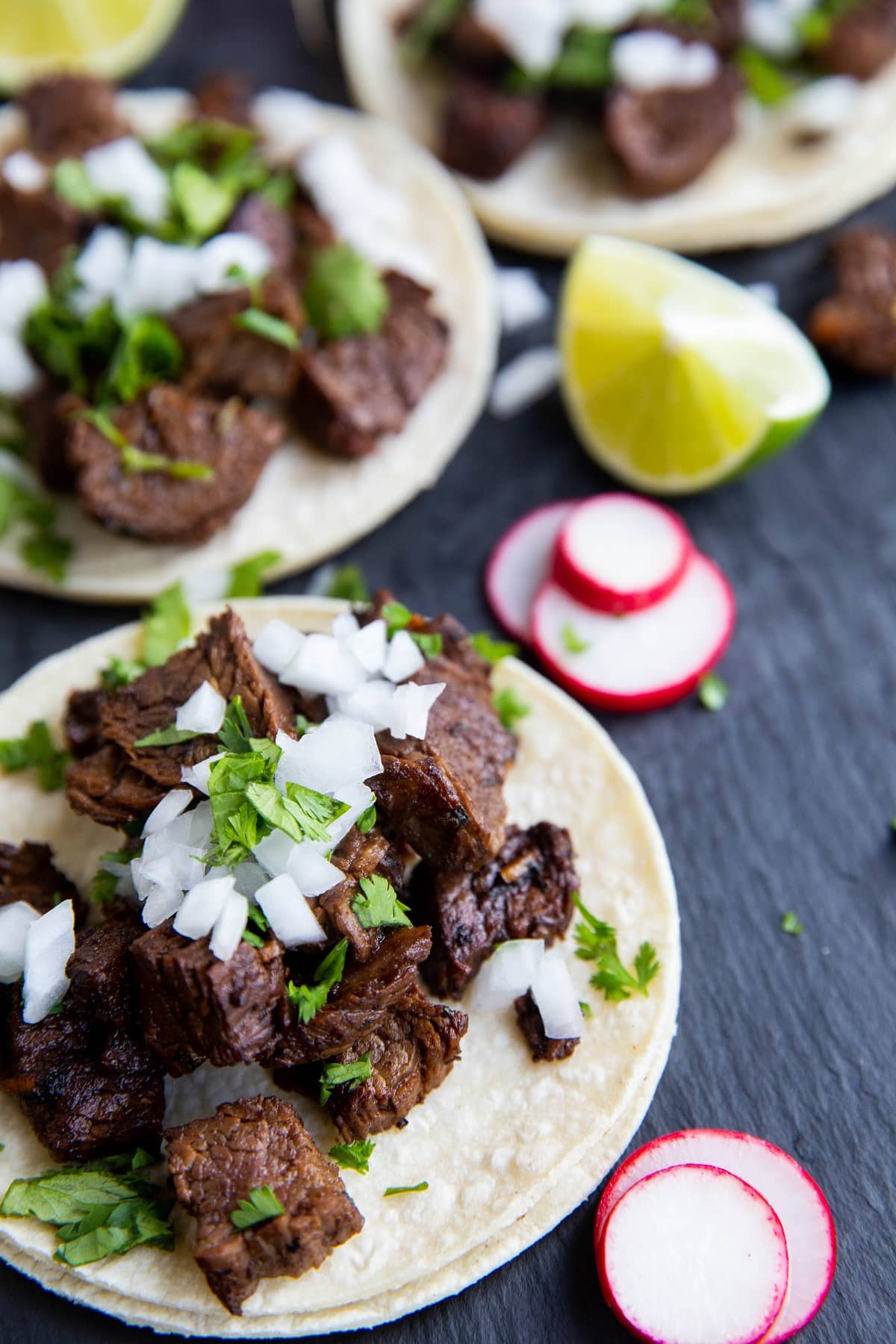 three carne asada tacos on a black slate with slices of turnips and limes to the sides