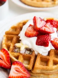 Stack of Belgian waffles on a white plate with whipped cream, butter, and strawberries