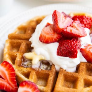 Stack of Belgian waffles on a white plate with whipped cream, butter, and strawberries