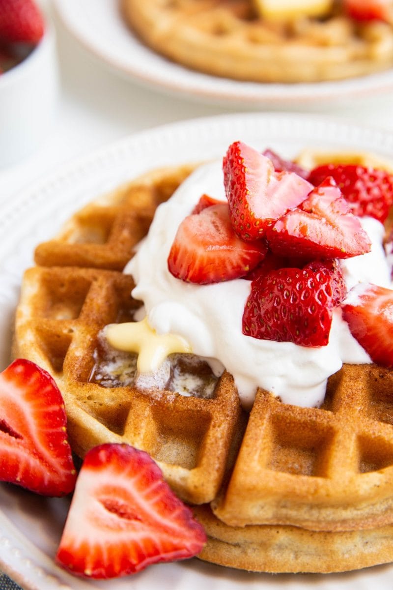 Stack of Belgian waffles on a white plate with whipped cream, butter, and strawberries