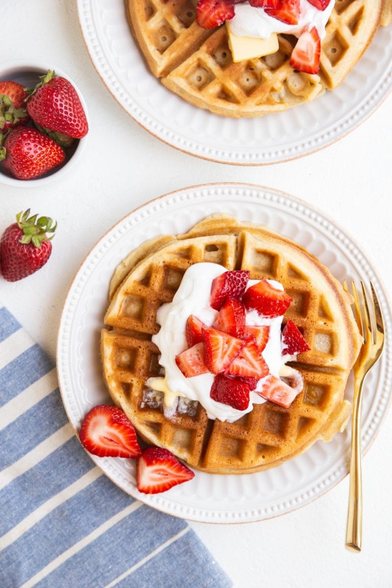 top down photo of two plates of waffles with whipped cream, strawberries, and a blue striped napkin