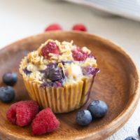 Berry baked oatmeal muffin on a wooden plate with fresh berries all around and more muffins in the background