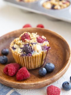 Berry baked oatmeal muffin on a wooden plate with fresh berries all around and more muffins in the background