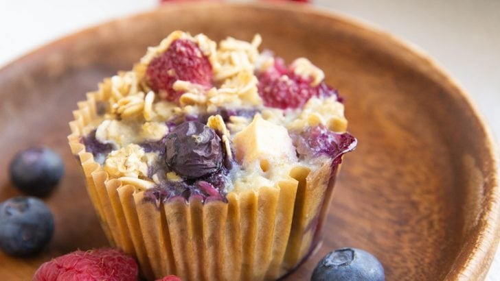 Berry baked oatmeal muffin on a wooden plate with fresh berries all around and more muffins in the background