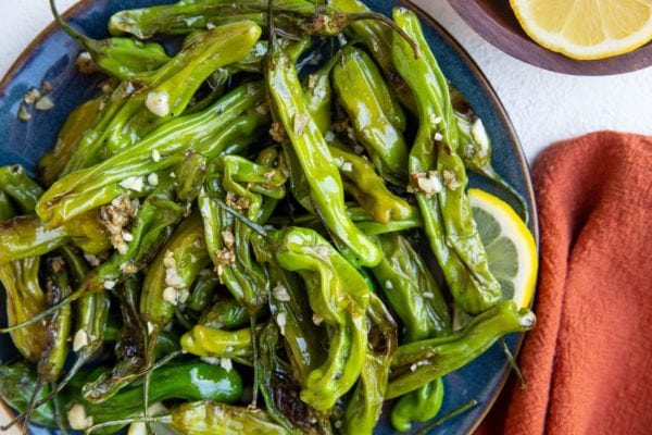 blue plate with cooked shishito peppers with a red napkin to the side and lemon slices.