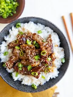 Black bowl with white rice and Korean beef sprinkled with sesame seeds and green onions. Chop sticks to the side, a bowl of green onions and a golden napkin
