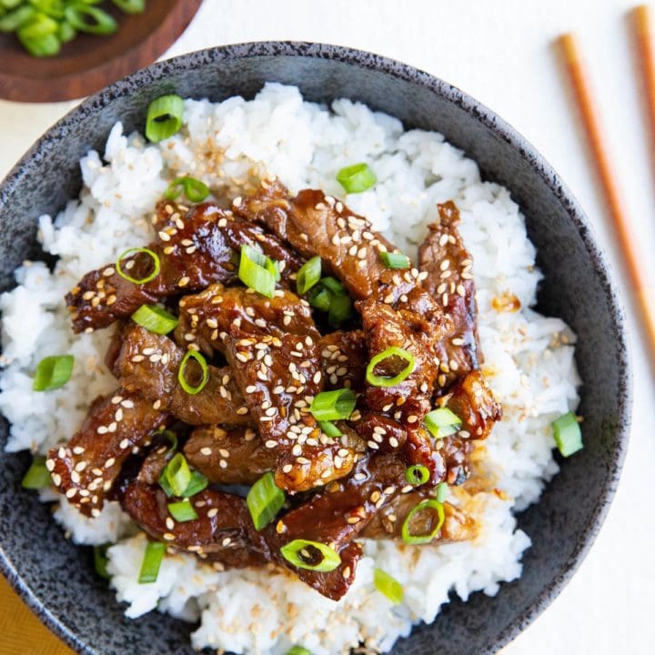 Black bowl with white rice and Korean beef sprinkled with sesame seeds and green onions. Chop sticks to the side, a bowl of green onions and a golden napkin