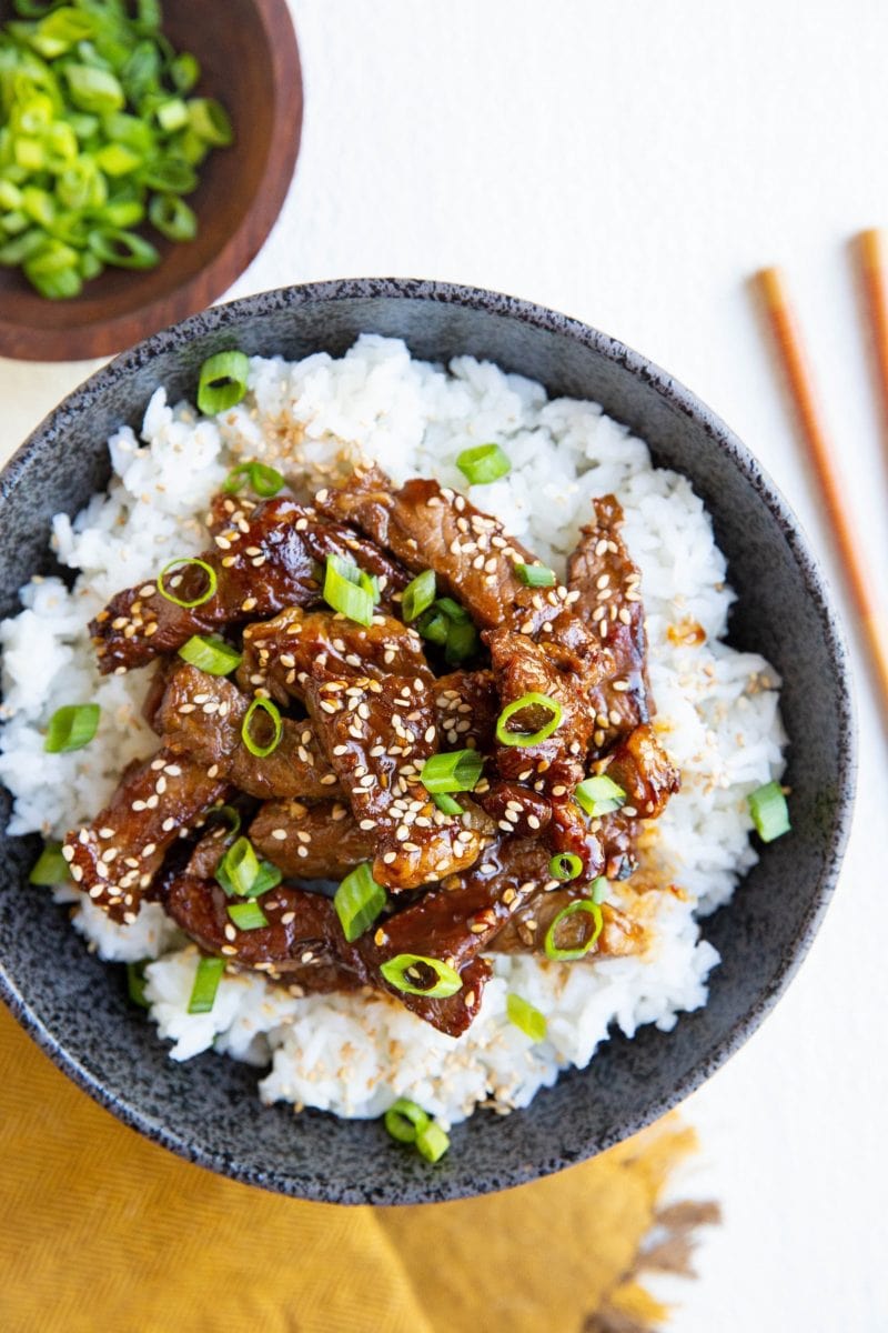 Black bowl with white rice and Korean beef sprinkled with sesame seeds and green onions. Chop sticks to the side, a bowl of green onions and a golden napkin