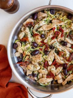 Stainless steel skillet with creamy sun-dried tomato chicken and zoodles with a red napkin to the side and a pepper shaker