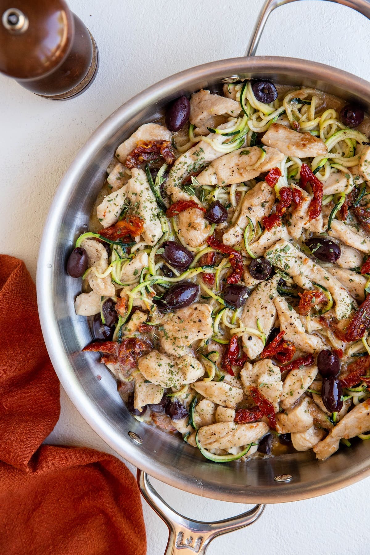 Stainless steel skillet with creamy sun-dried tomato chicken and zoodles with a red napkin to the side and a pepper shaker