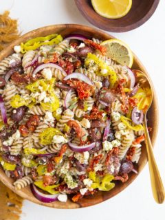 Wooden bowl of Greek pasta salad with a gold spoon, bowl of lemons and golden napkin