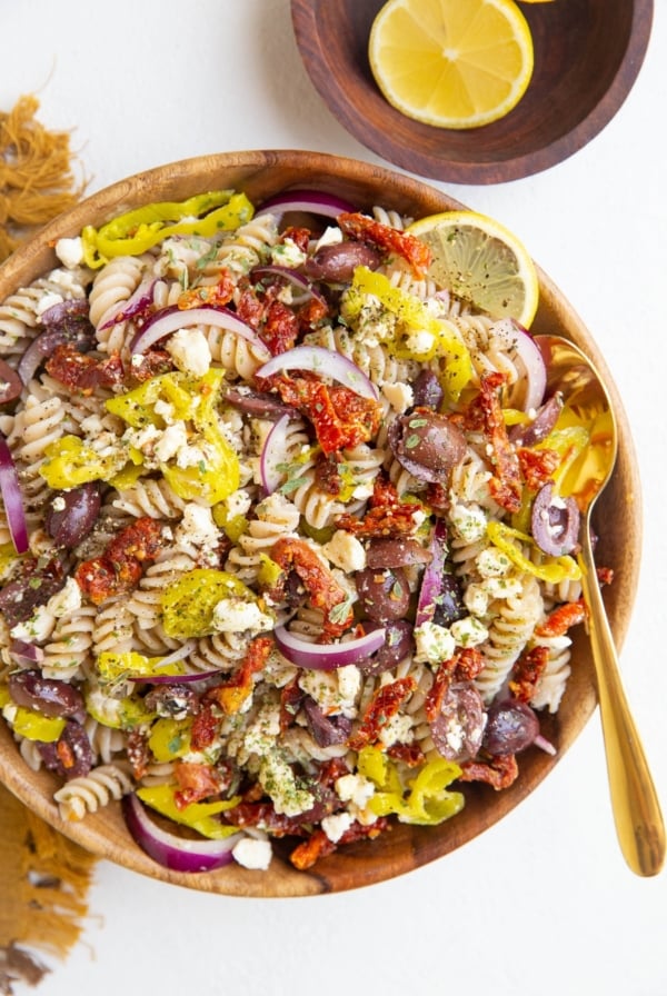 Wooden bowl of Greek pasta salad with a gold spoon, bowl of lemons and golden napkin