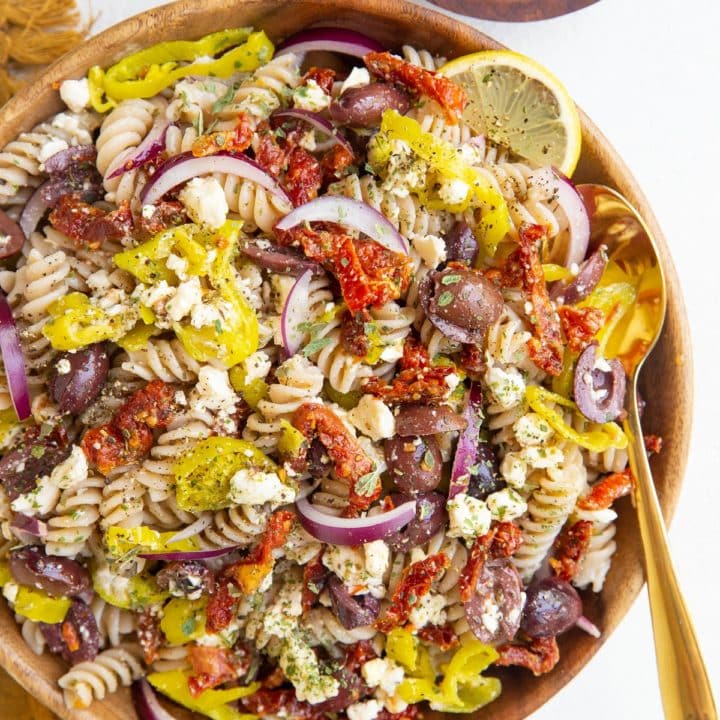 Wooden bowl of Greek pasta salad with a gold spoon, bowl of lemons and golden napkin