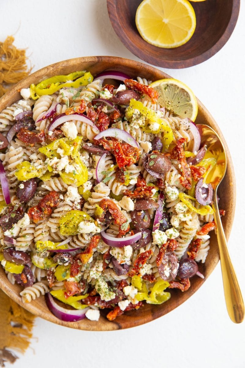 Wooden bowl of Greek pasta salad with a gold spoon, bowl of lemons and golden napkin
