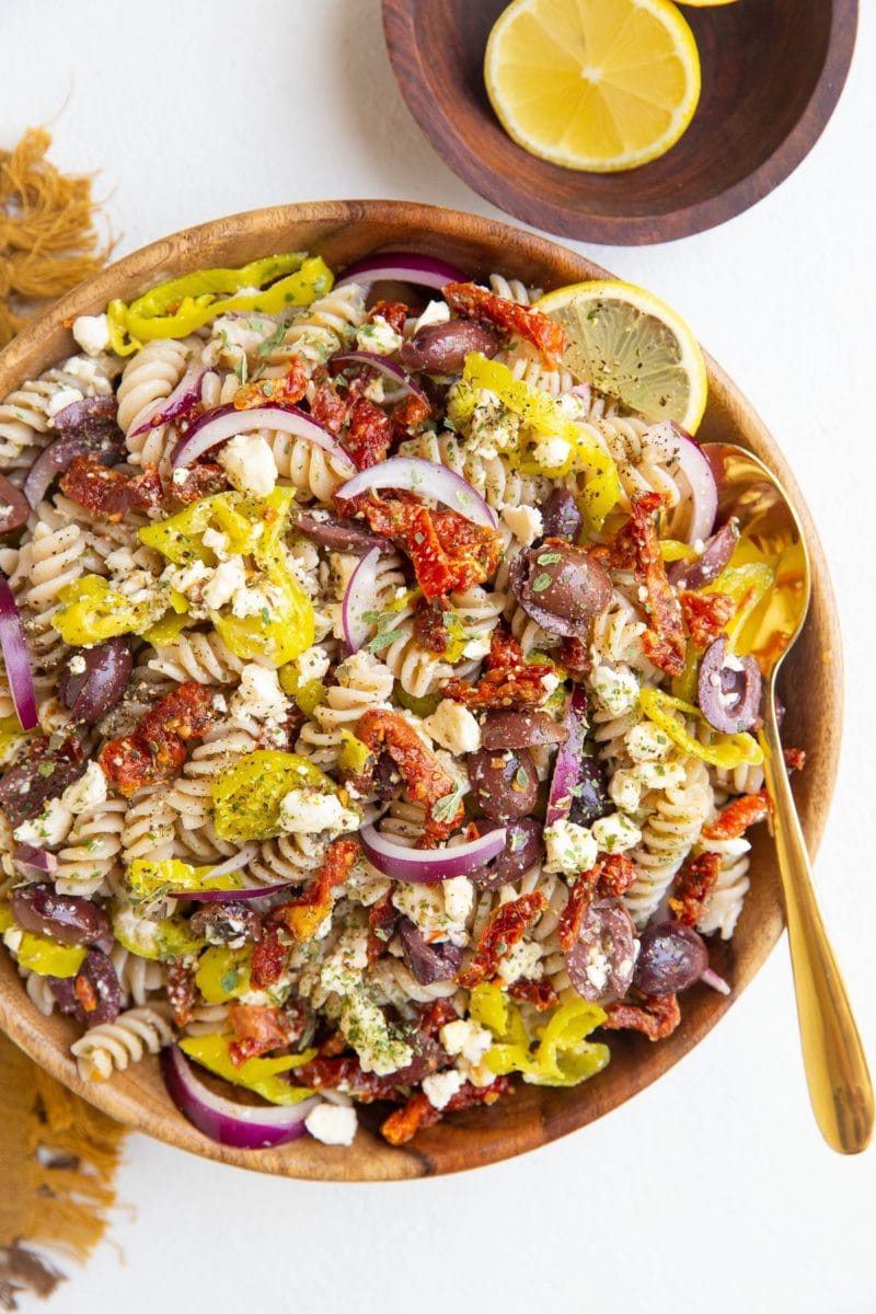Wooden bowl of Greek pasta salad with a gold spoon, bowl of lemons and golden napkin