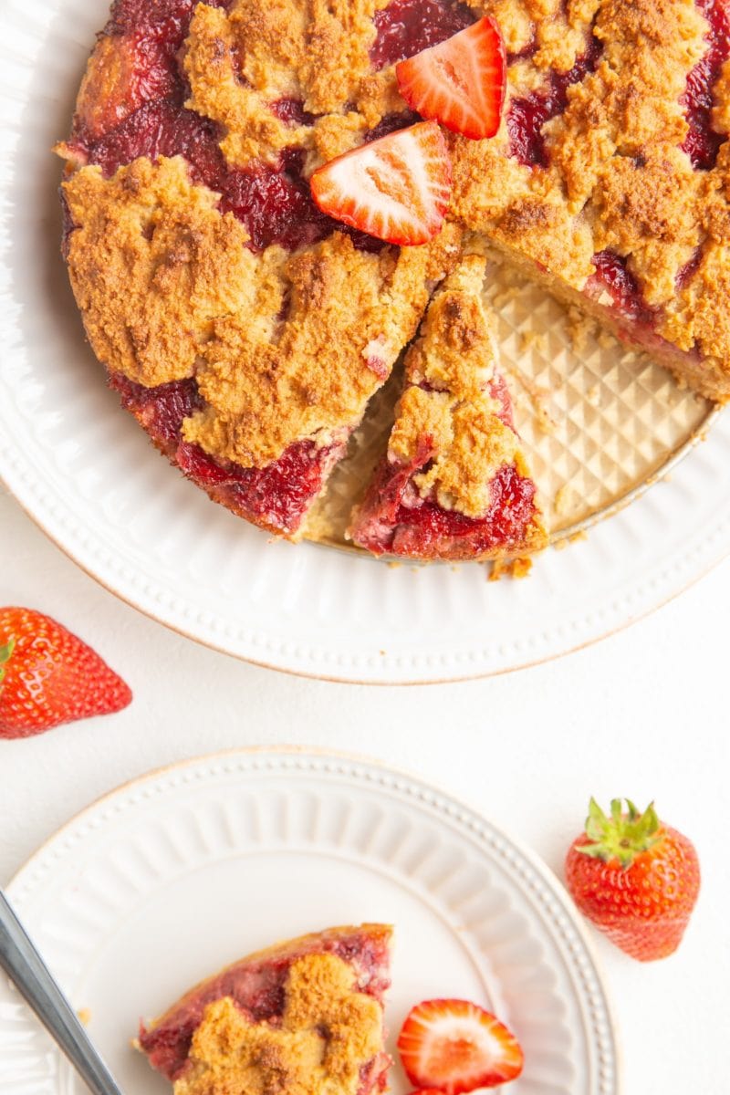 Top down photo of a sliced strawberry crumb cake with a slice of cake on a white plate
