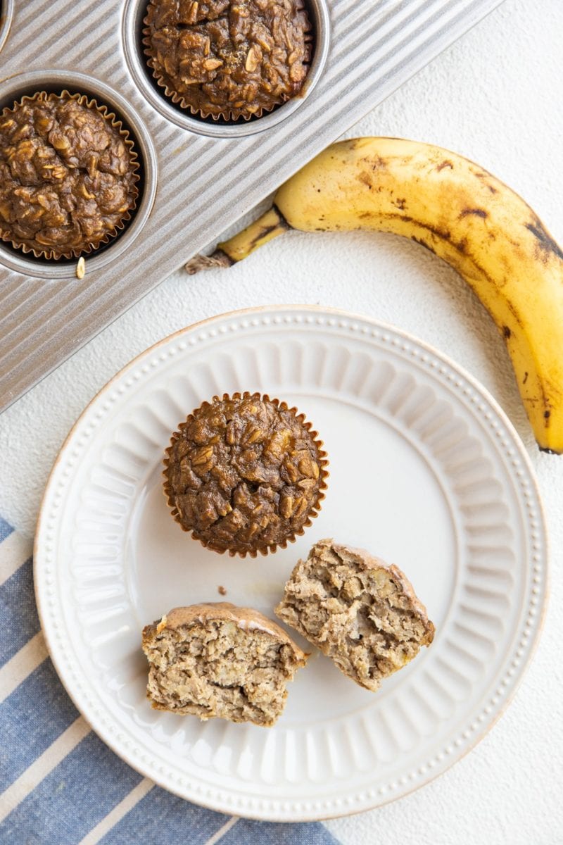 plate of baked oatmeal muffins and a muffin tray with baked muffins inside. A blue napkin to the side and a ripe banana