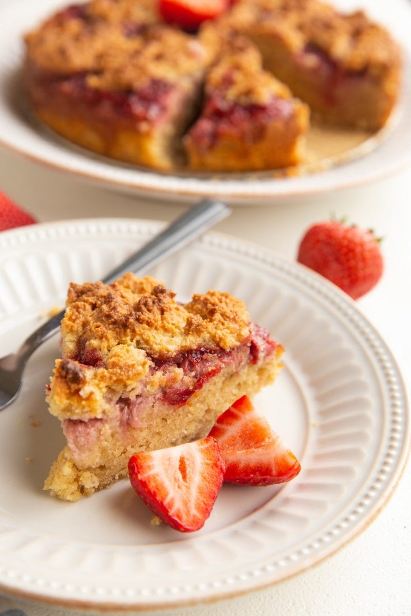 slice of strawberry crumb cake on a white plate with the rest of the cake in the backfround