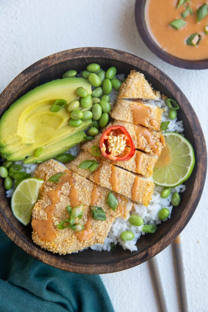 wooden bowl with rice, crispy chicken, avocado, edamame and a blue napkin and chopsticks to the side