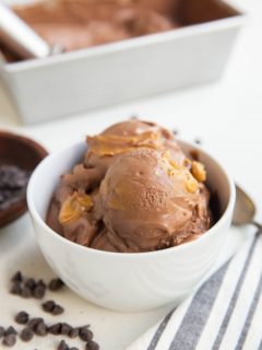 white bowl of several scoops of chocolate peanut butter ice cream with a pan of ice cream in the background, a bowl of chocolate chips and a spoon and napkin off to the side.