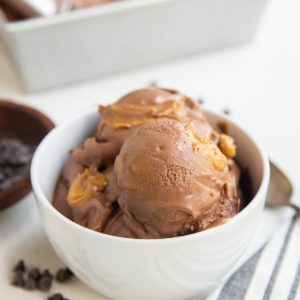 white bowl of several scoops of chocolate peanut butter ice cream with a pan of ice cream in the background, a bowl of chocolate chips and a spoon and napkin off to the side.