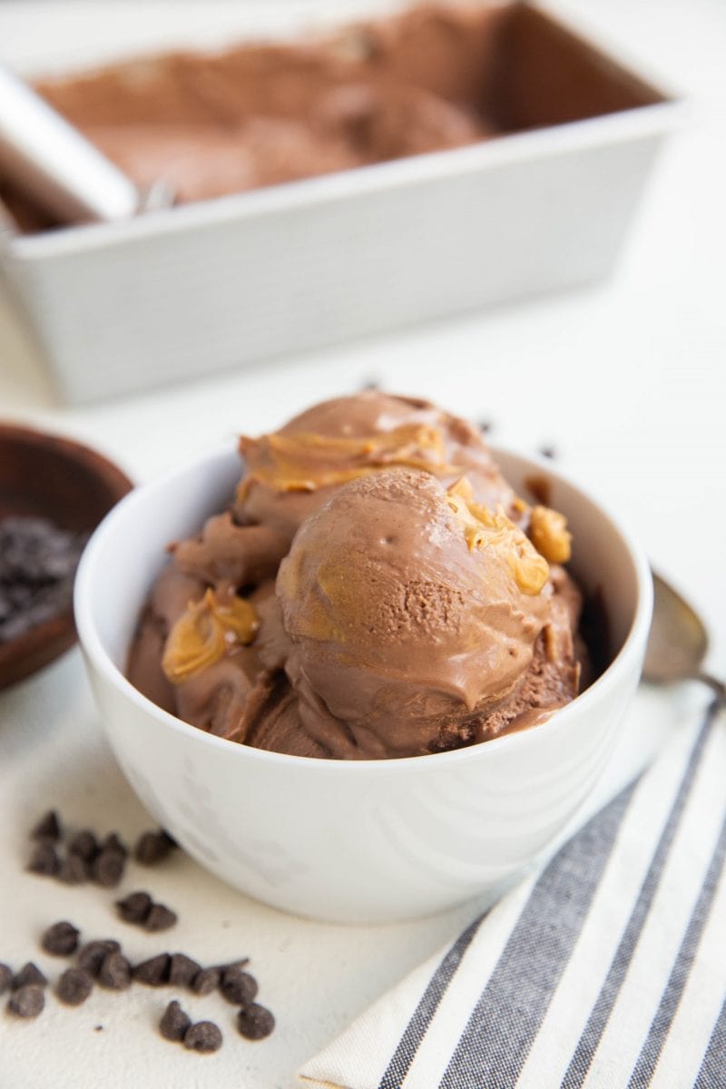 white bowl of several scoops of chocolate peanut butter ice cream with a pan of ice cream in the background, a bowl of chocolate chips and a spoon and napkin off to the side.