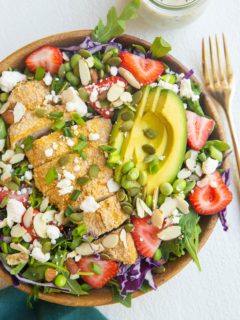 bowl of crispy chicken salad with strawberries. blue napkin and gold fork off to the side.