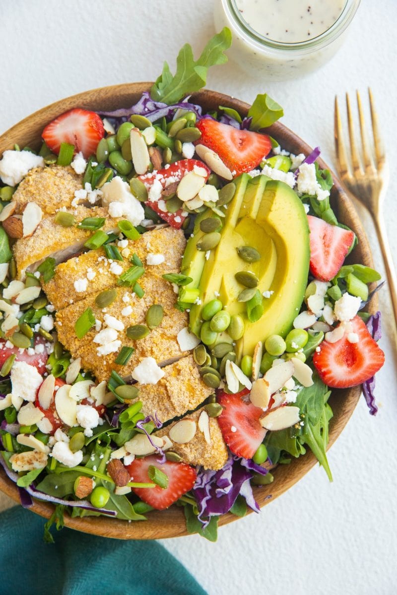 bowl of crispy chicken salad with strawberries. blue napkin and gold fork off to the side.